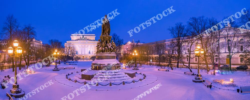 На фотографии изображена площадь Островского в зимнее время. Центральное место занимает памятник Екатерине II, окруженный заснеженными аллеями и фонарями, создающими уютную атмосферу. Величественный Александринский театр подсвечен мягким светом, добавляя торжественности пейзажу. Глубокий синий оттенок вечернего неба подчеркивает красоту зимнего Санкт-Петербурга.